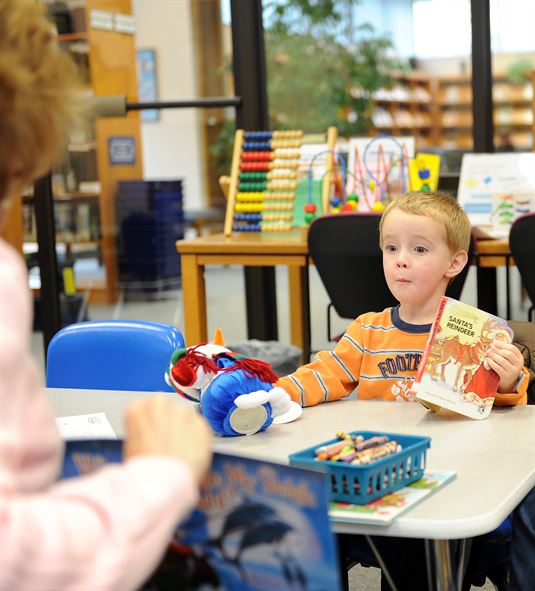 A child holds a book and toy while watching an adult read a book.