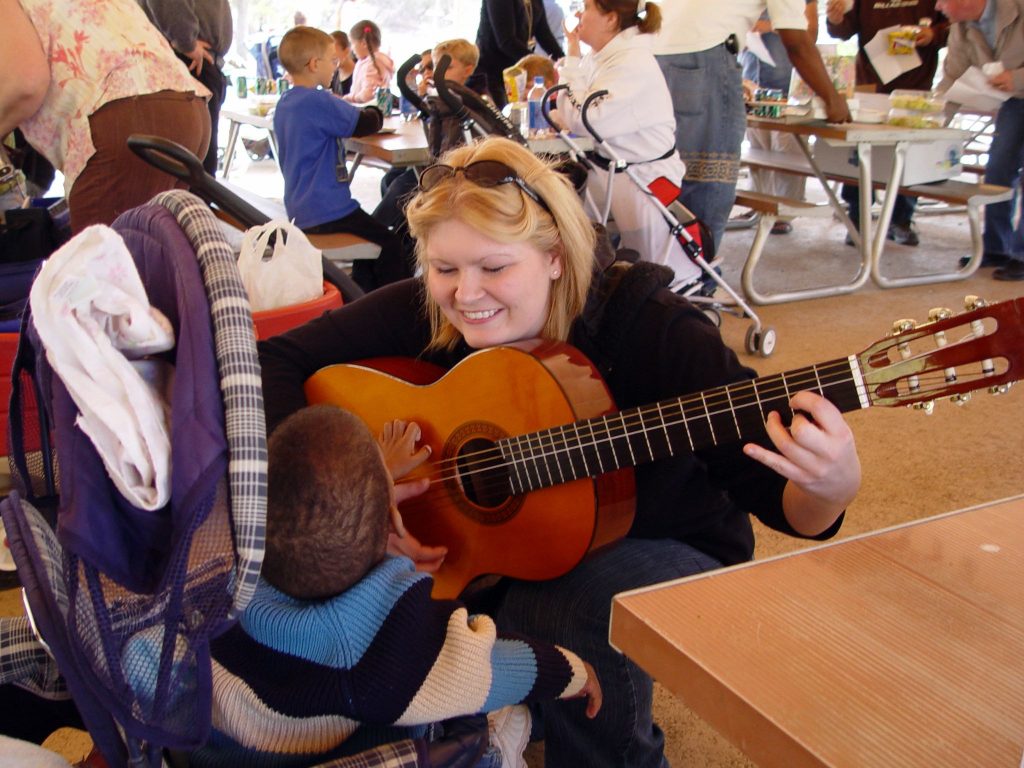 Photo of a woman holding a guitar, smiling at a child who is touching the guitar. Other people are shown in the background.