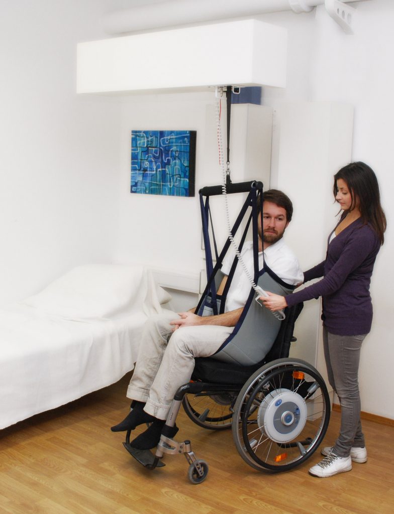 Photo showing a simulated patient and healthcare worker using a patient lift to move patient between bed and wheelchair