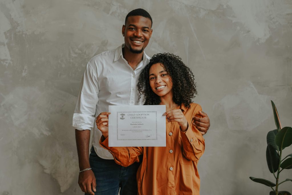 Photo showing a man and woman smiling at the viewer, the woman holding an adoption certificate