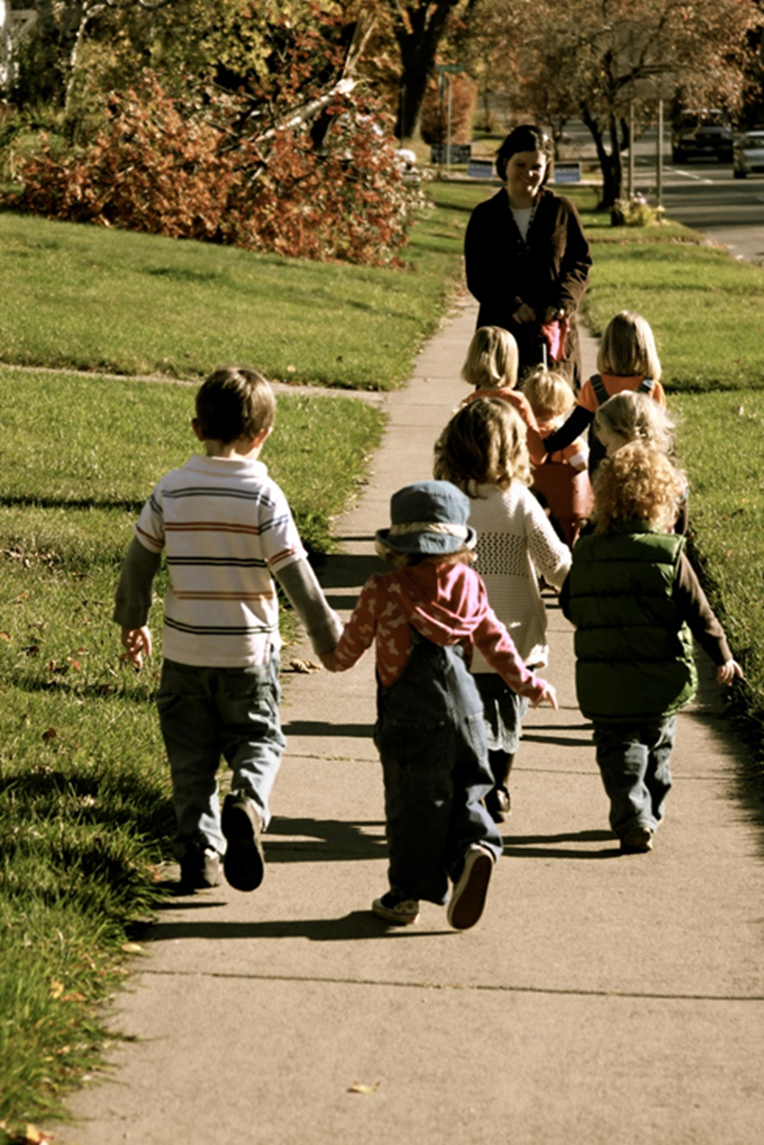 “IMG_2791” by Nora Zubriski is licensed under CC BY-NC 4.0 Image showing a childcare provider walking down a sidewalk with a small group of children