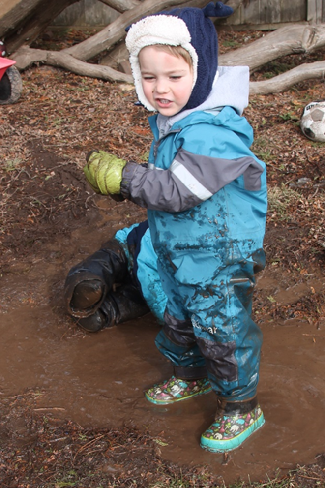 ”IMG-0835” by Nora Zubriski is licensed under CC BY-NC 4.0 Image showing a small, warmly dressed child standing in a mud puddle, looking happy to be covered in mud