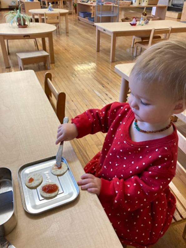 “IMG_6958” by Nora Ziburski is licensed under CC BY-NC 4.0 Image showing a child standing in a Montessori toddler classroom, spreading jam on a biscuit with a small knife