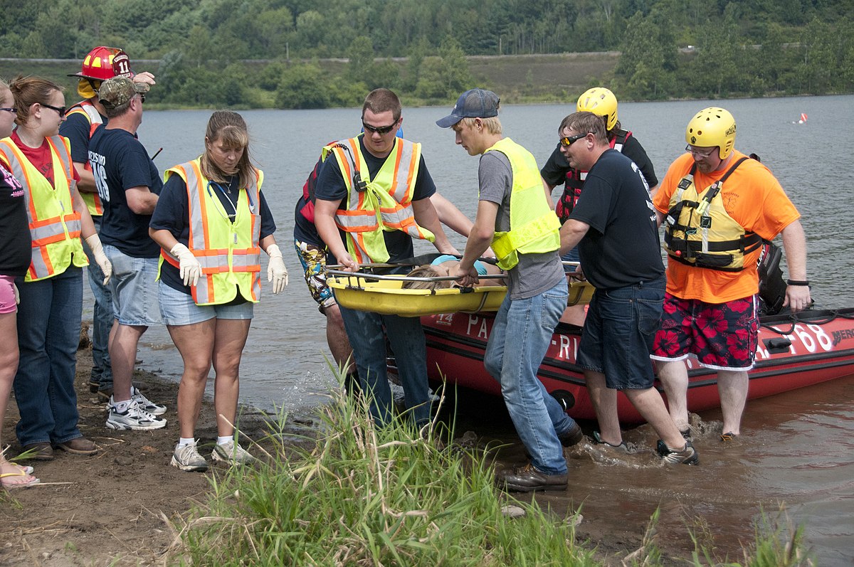 A group of rescuers and emergency personnel are on the shore, performing a simulated water rescue